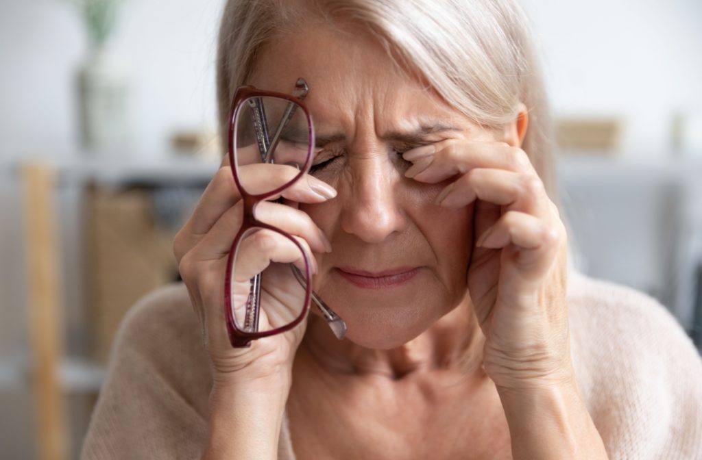 Older woman rubbing her eyes while holding her glasses, looking distressed and showing signs of dealing with early symptoms of aging eyes.