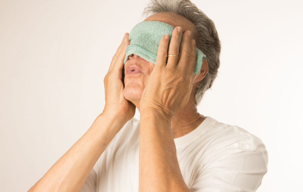 A person wearing a white shirt sighs in relief as they use a warm compress on their eyes to reduce eye pressure.