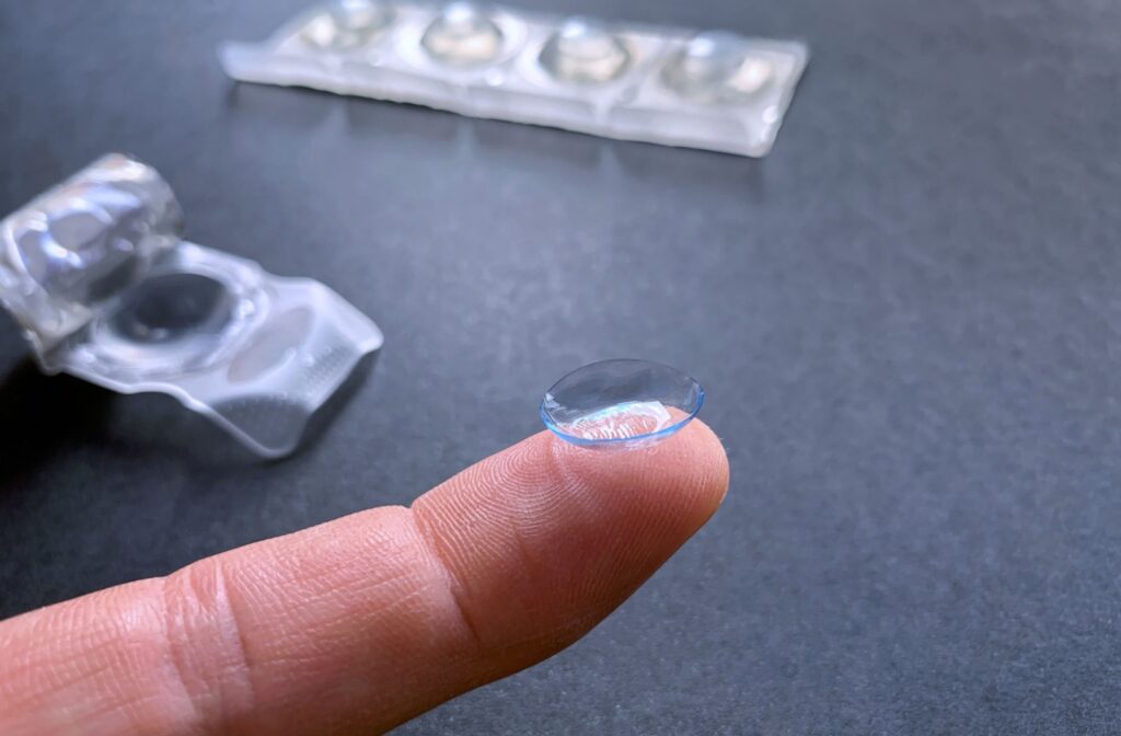A finger holds a dry contact lens that has expired. Contact lens blister packs appear in the background.